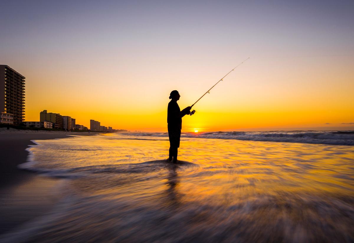 An angler silhouetted in the surf at sunset, with beachfront buildings behind