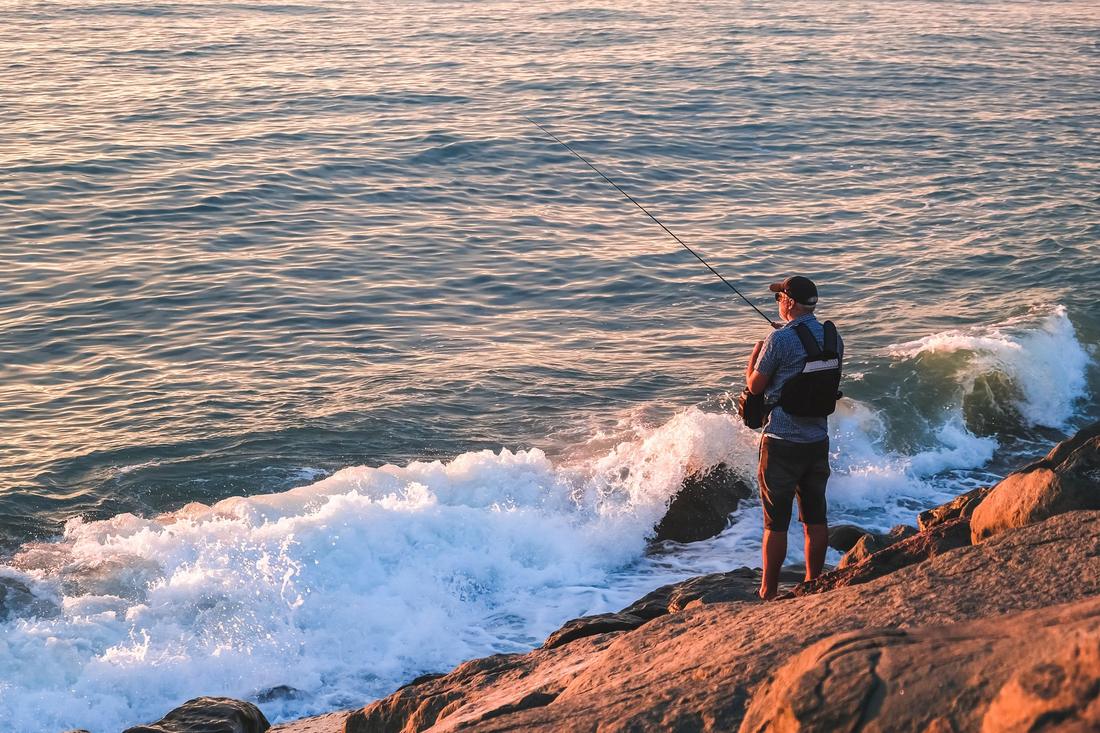 An angler fishing from rocks at sunrise as waves break
